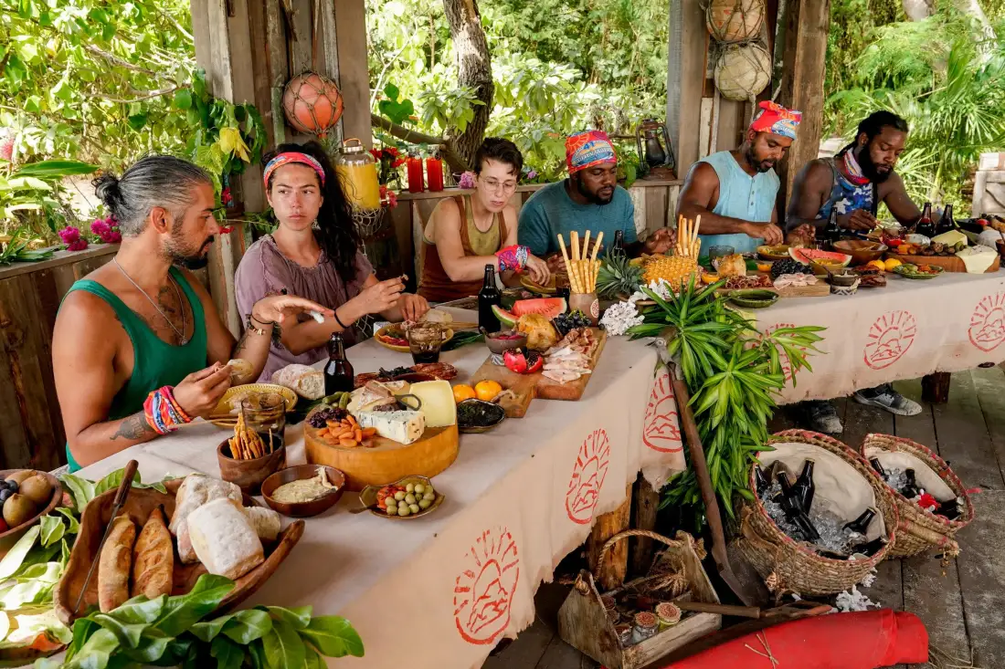 The merged Survivor 41 castaways eating at a long table laid with a merge feast